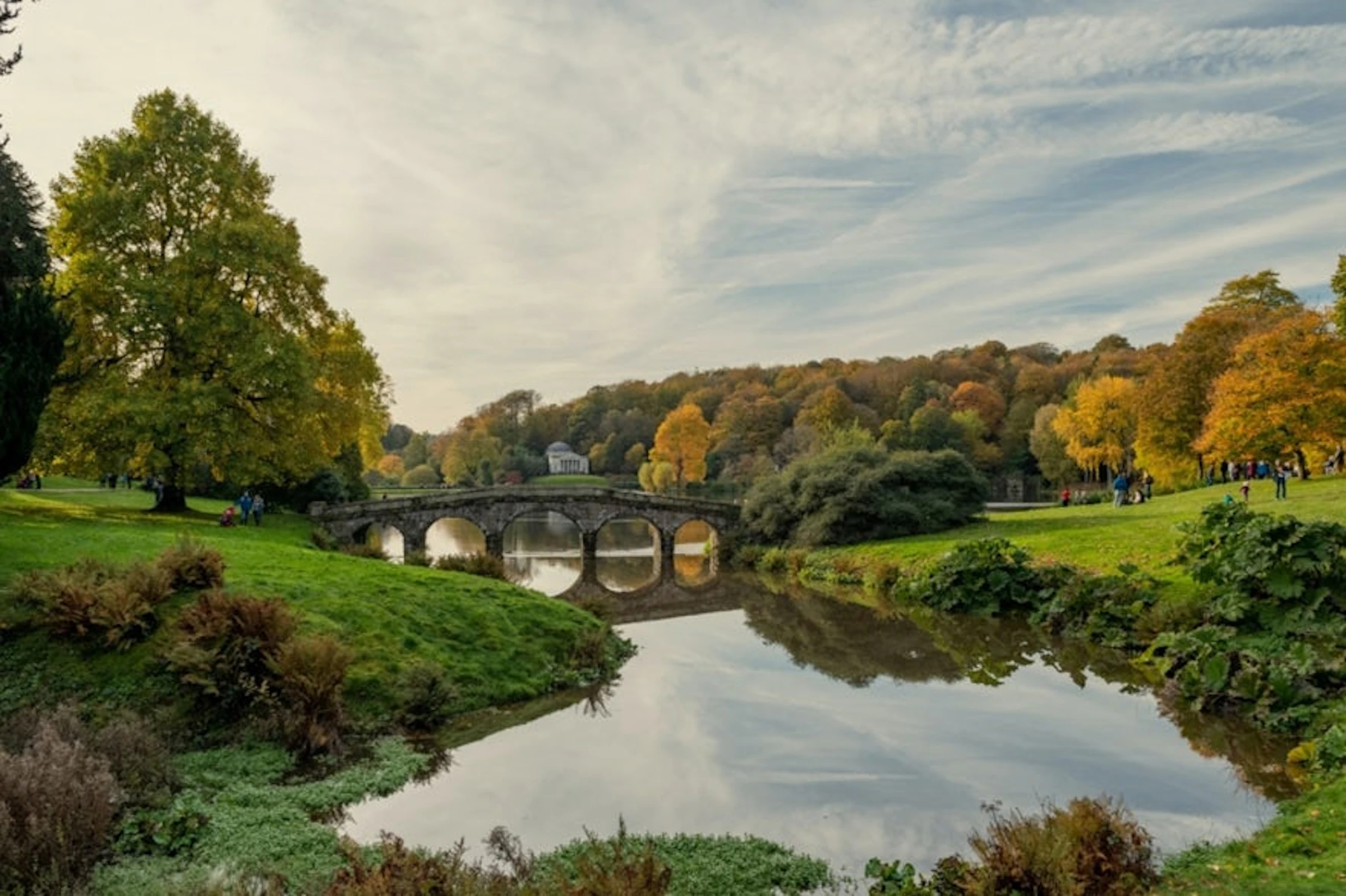 Stone bridge with arches over a calm river, surrounded by trees with autumn foliage under a partly cloudy sky.
