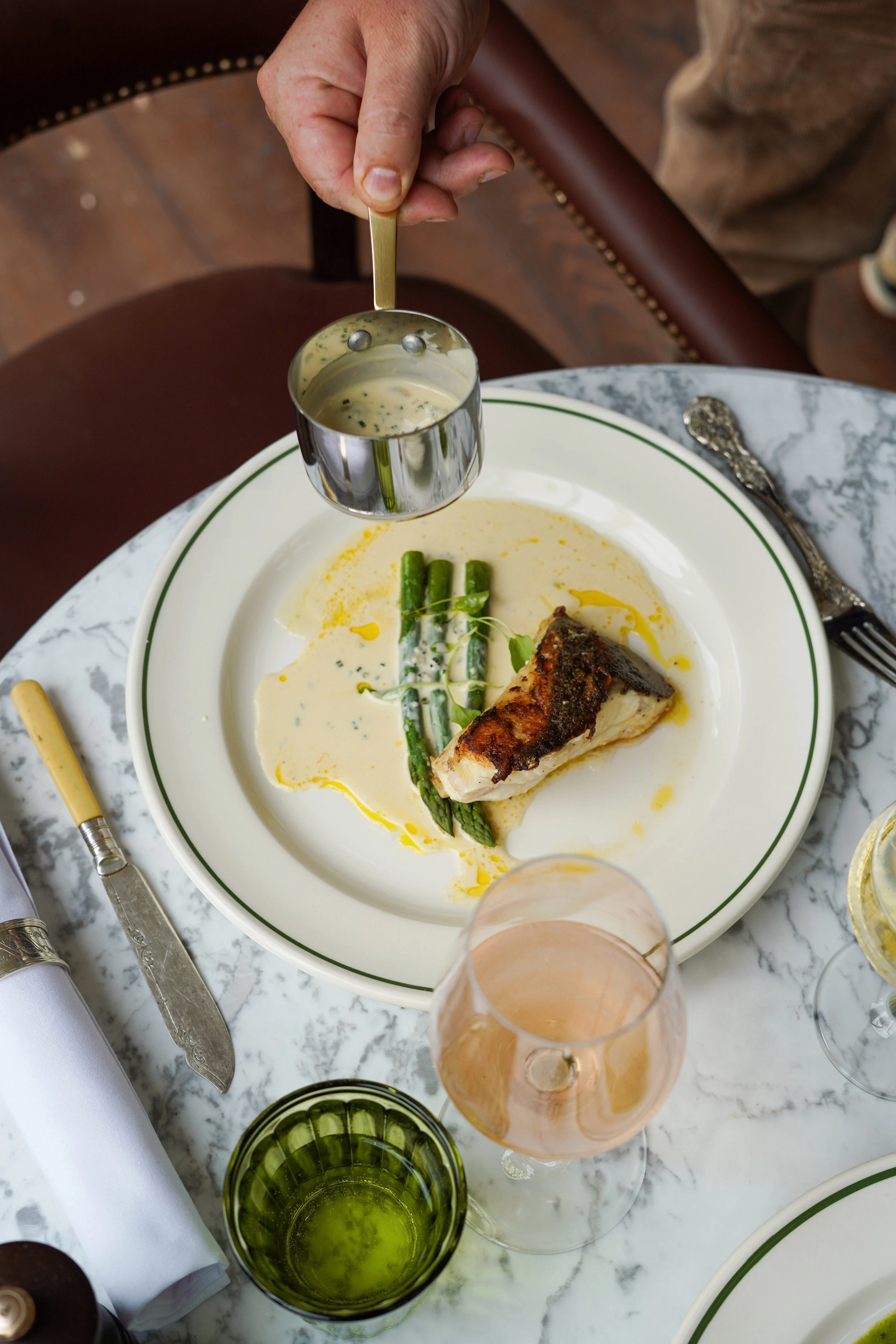 A hand pours sauce over grilled fish and asparagus on a white plate set on a marble table with drinks and cutlery.