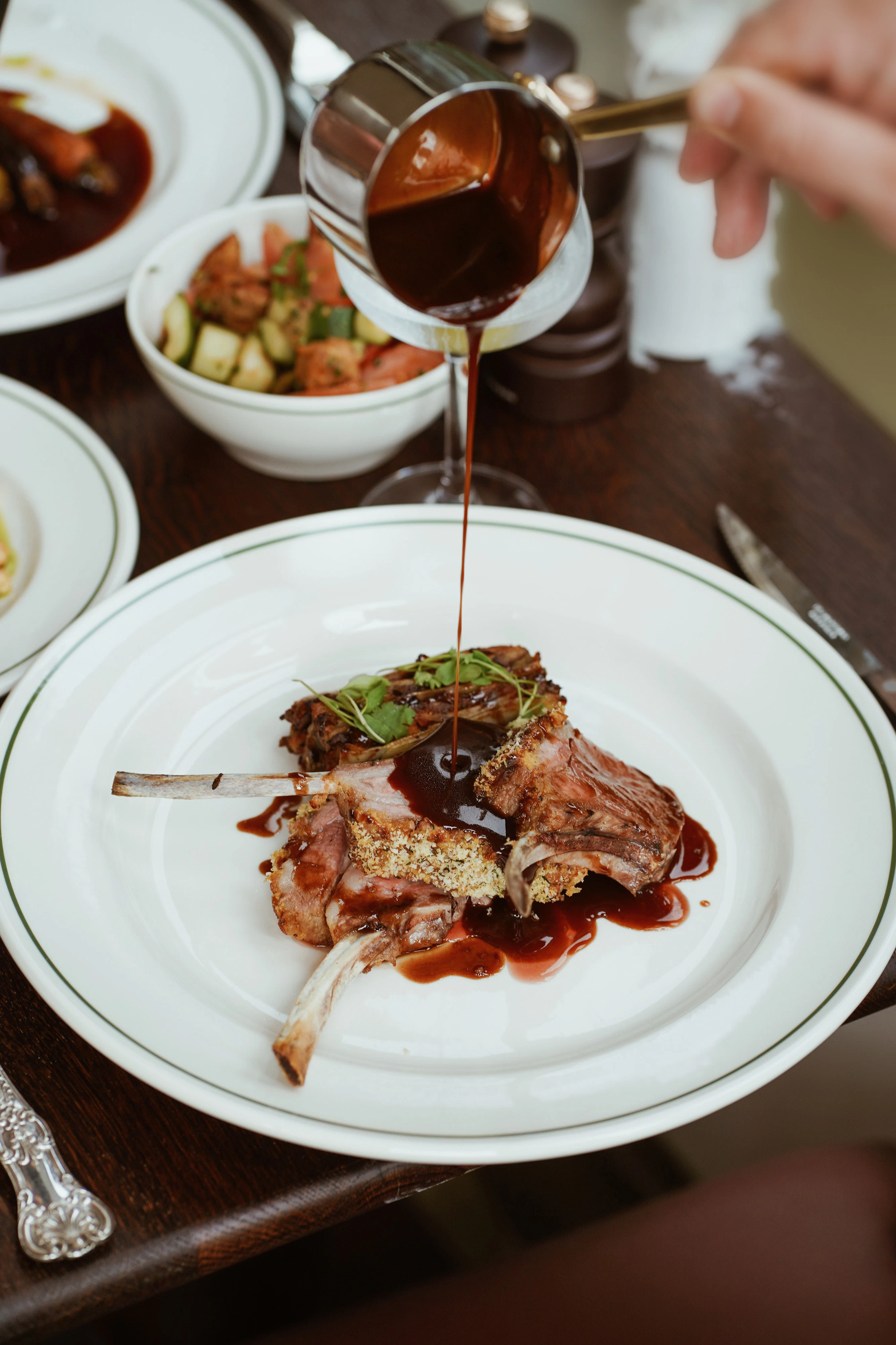 A person pours sauce over a plated lamb chop served with greens and vegetables on a white dish.
