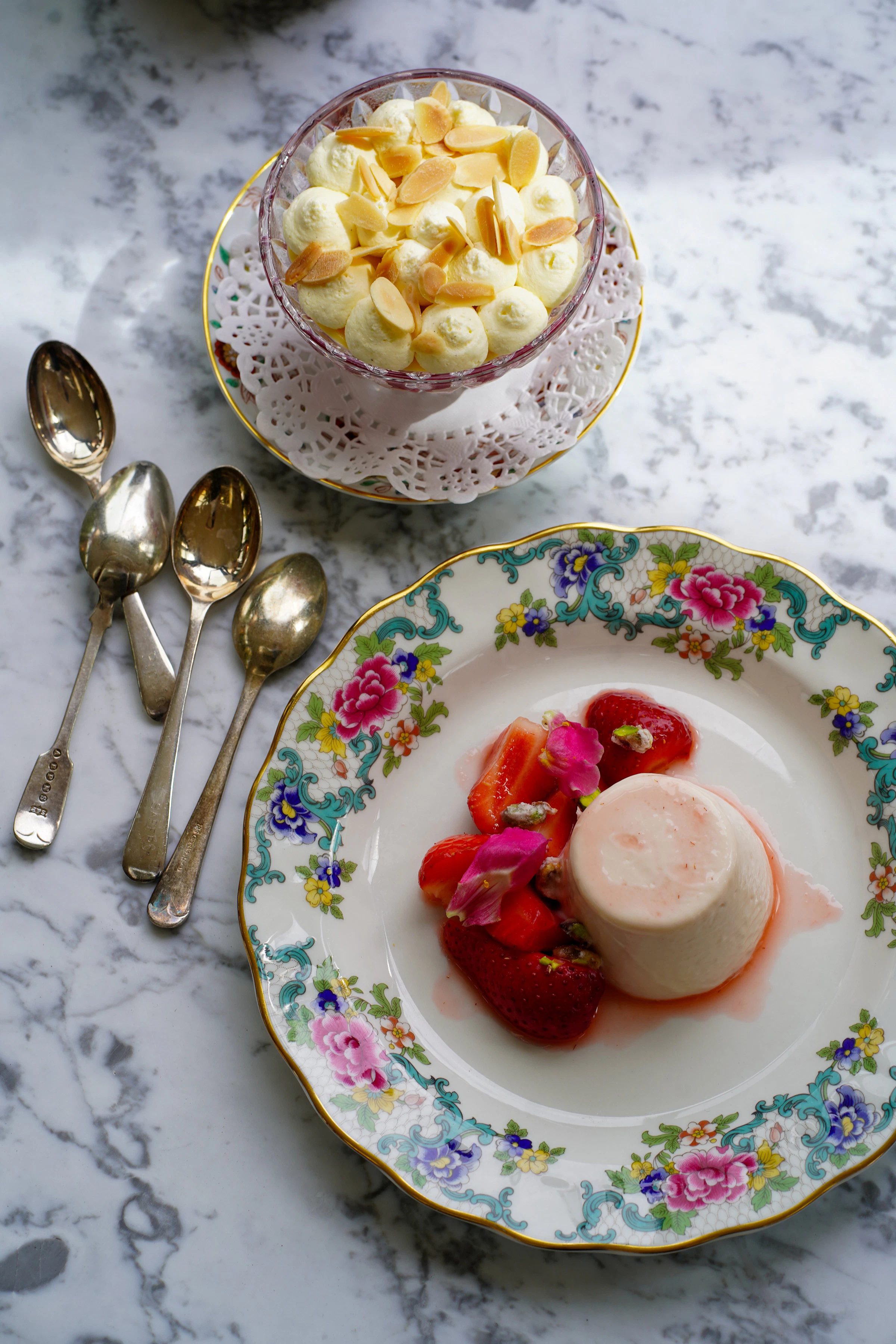 Dessert plate with panna cotta, strawberries, and flower petals beside a dish of cream with almonds and four spoons on marble.