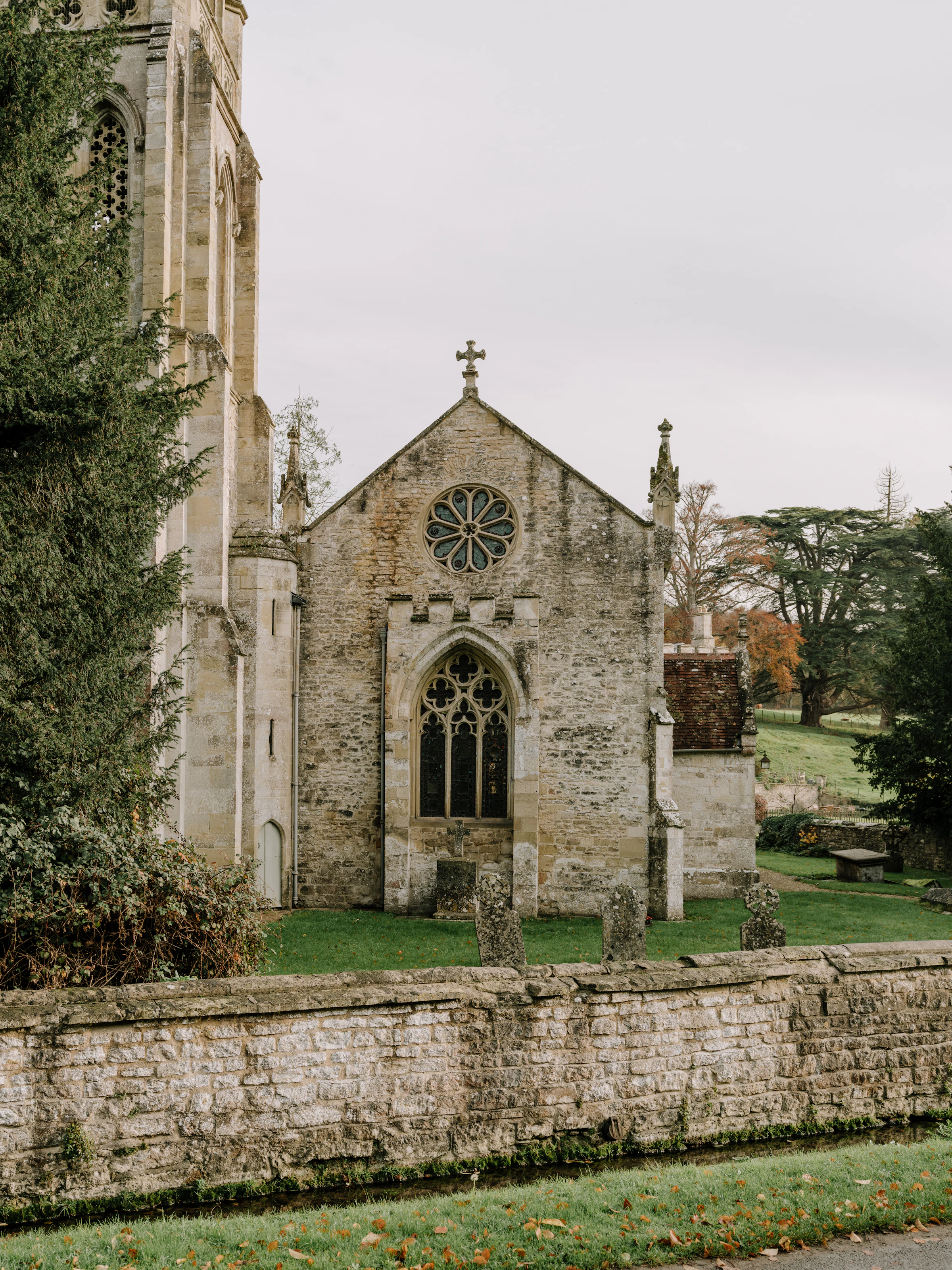 Stone church with arched windows and a rose window, surrounded by grass, trees, and a stone wall.