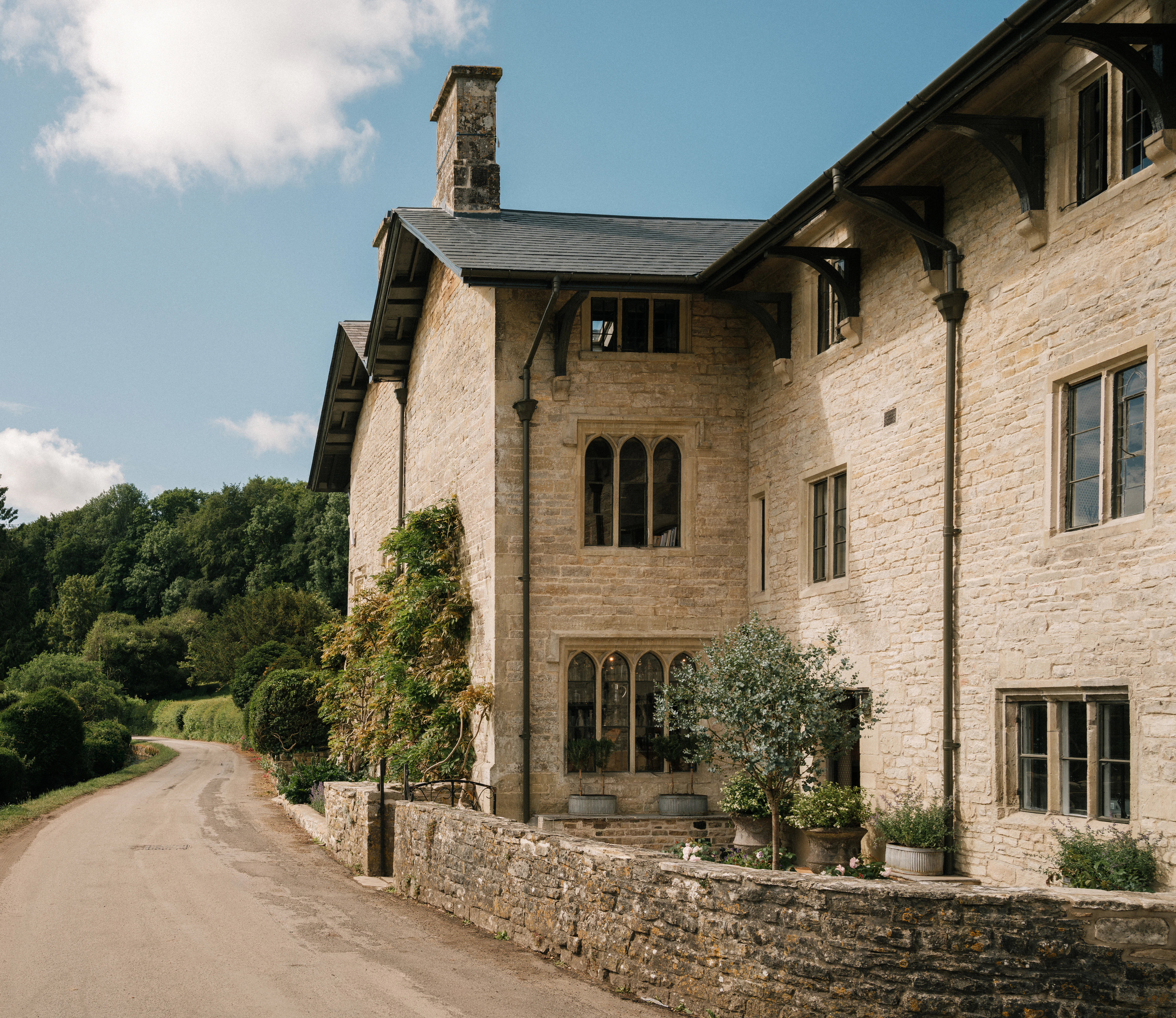 Stone house with arched windows and potted plants beside a quiet country road, surrounded by greenery.