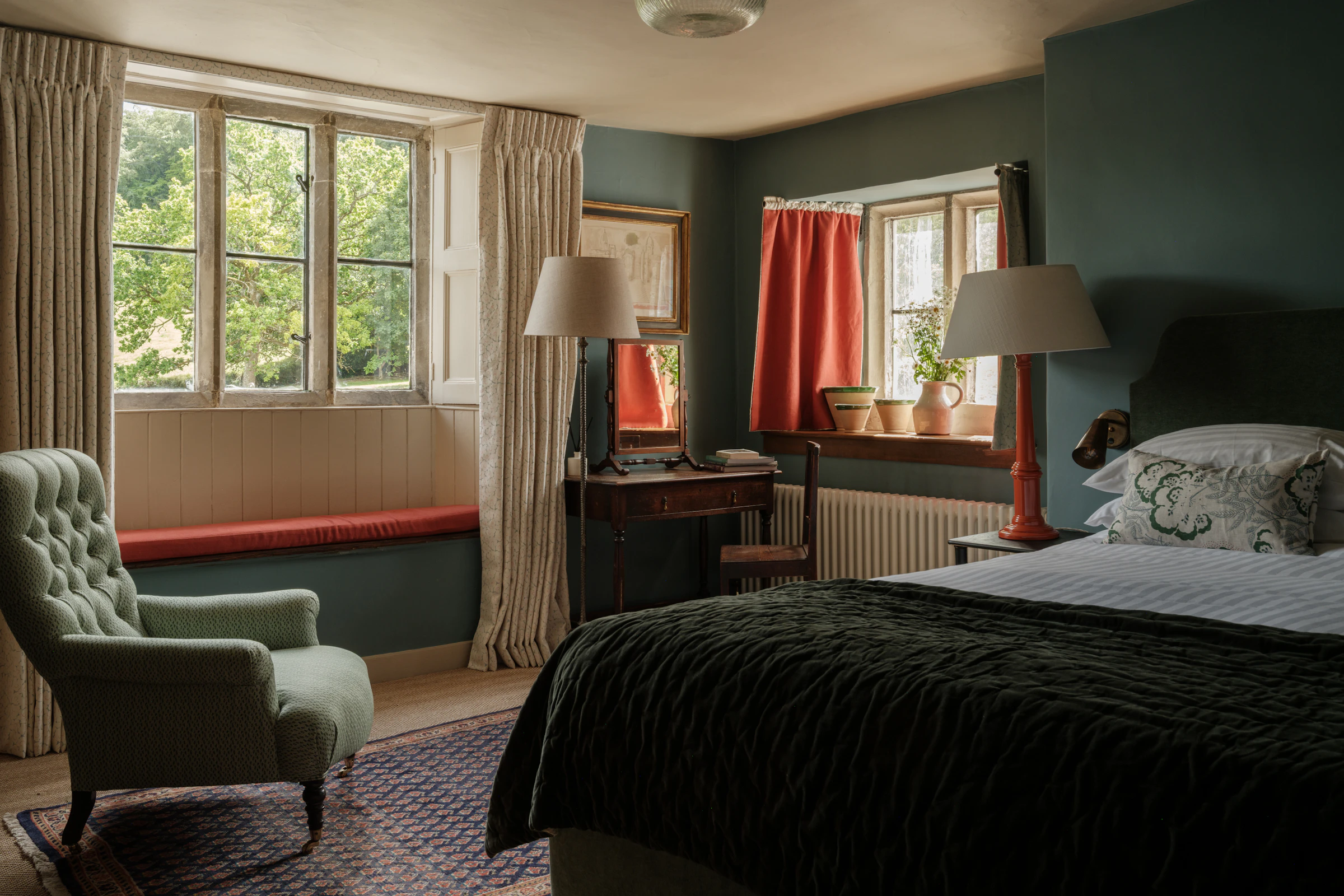 Cozy bedroom with armchair, bed, desk, lamps, plants, and large windows with curtains letting in natural light.