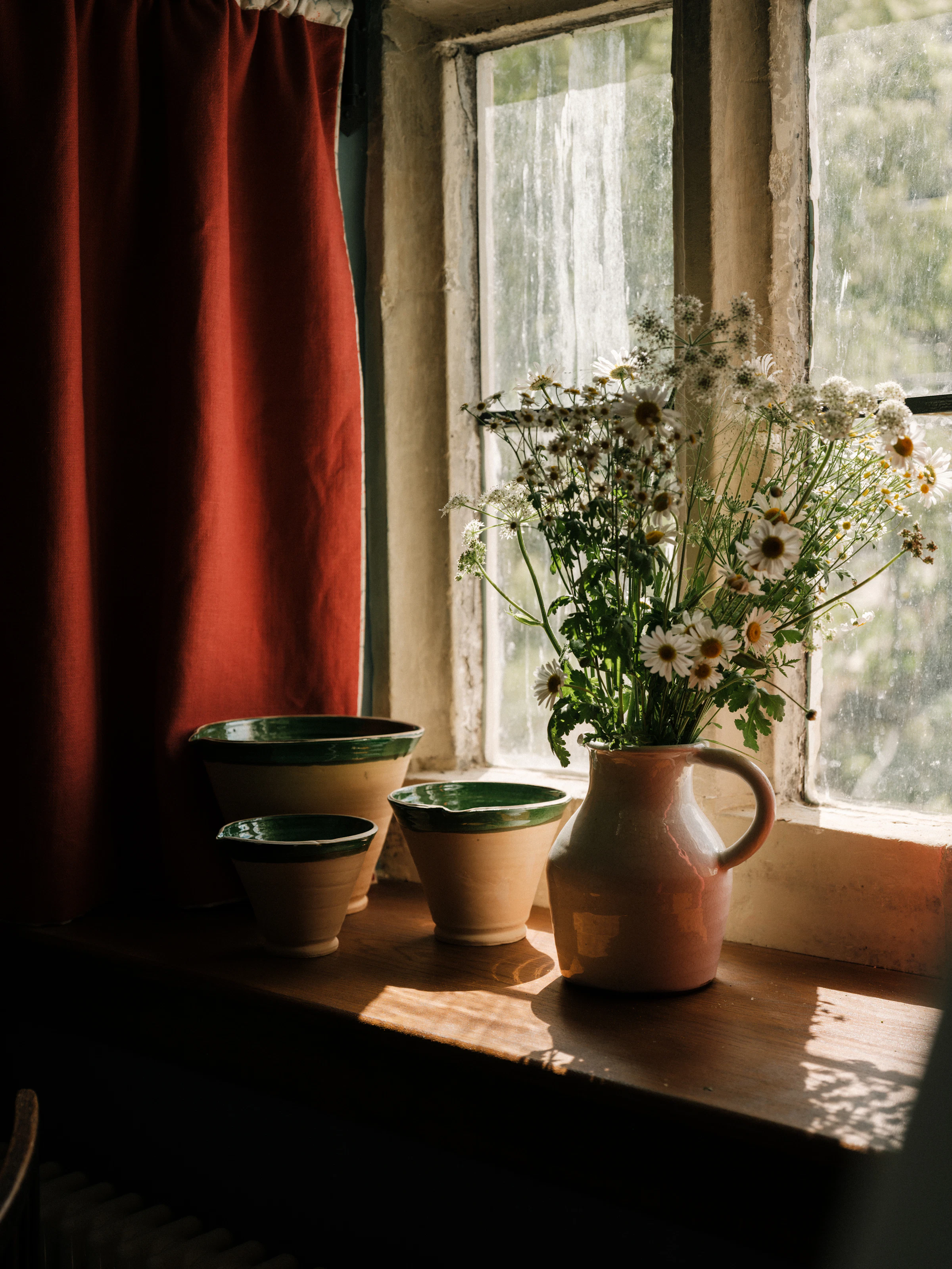 A ceramic jug with daisies and three bowls sit on a sunlit windowsill beside a red curtain.