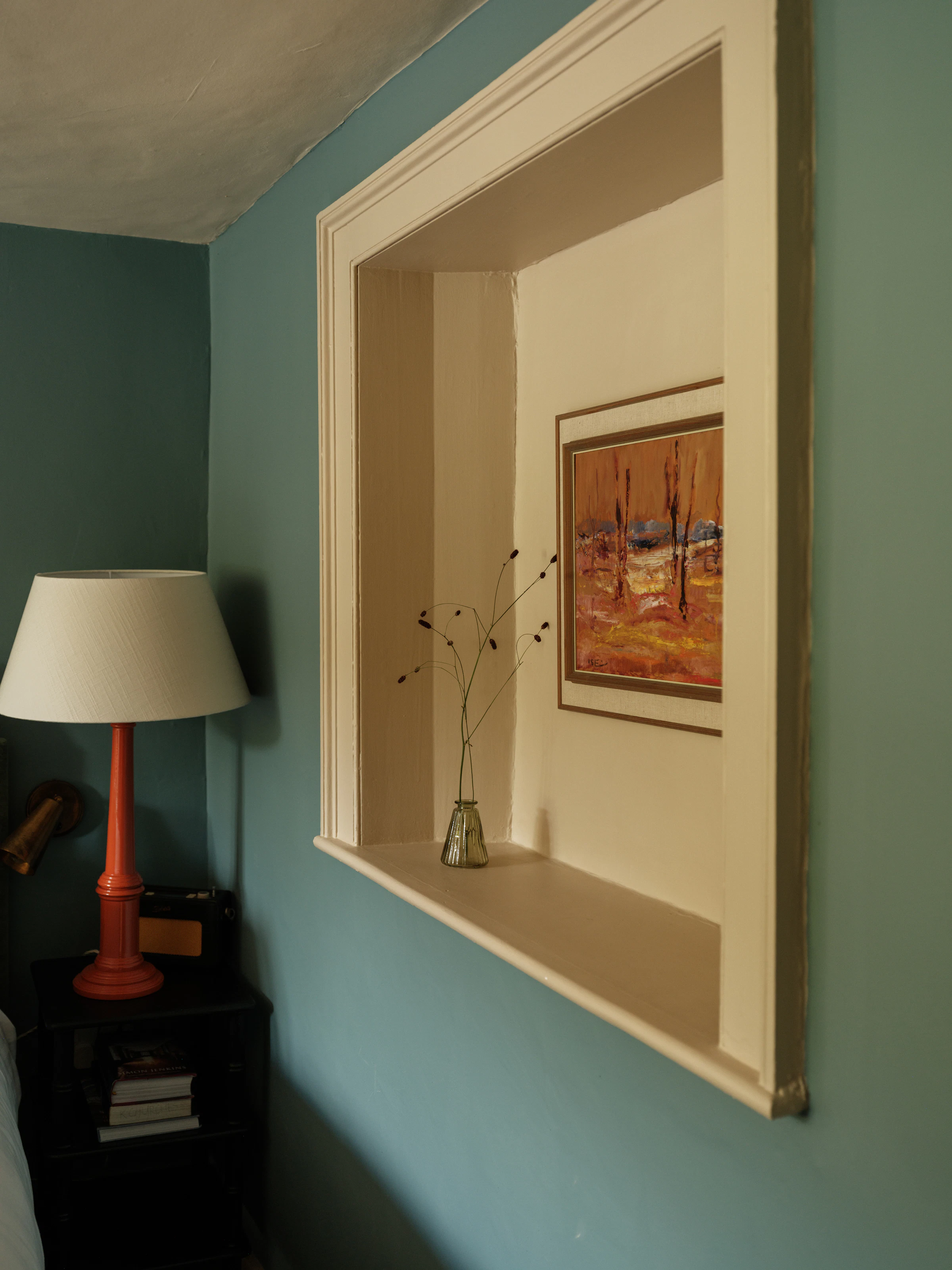 A small glass vase with dried flowers sits on a built-in shelf, below a framed landscape painting on a teal wall.