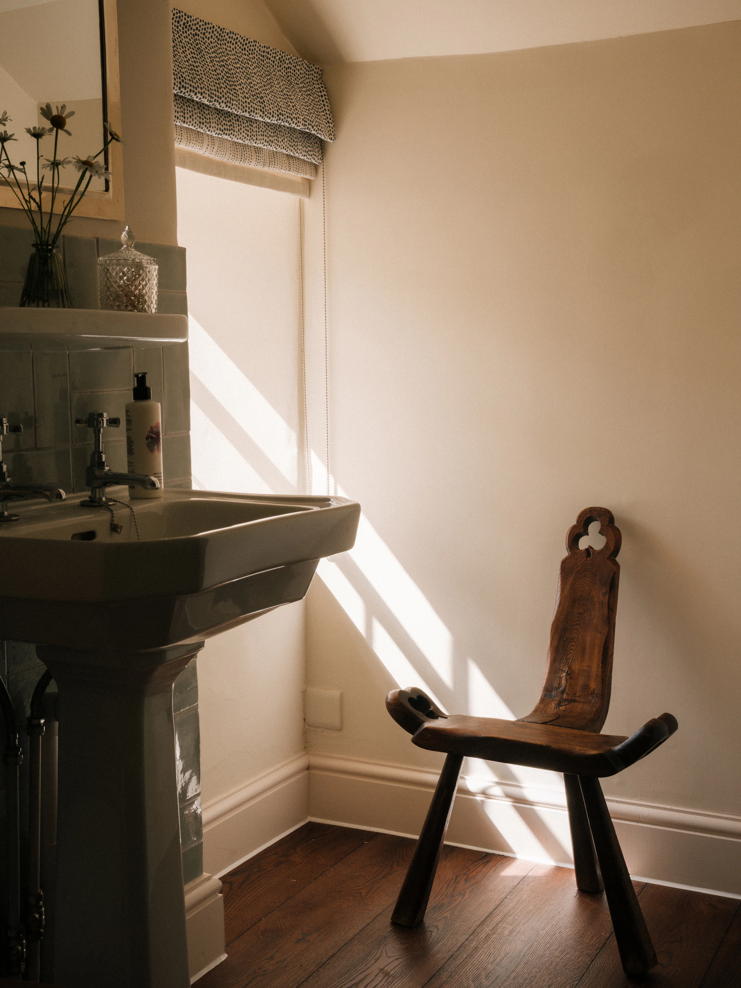 Sunlight streams through a window onto a rustic wooden chair and sink in a minimalist bathroom.