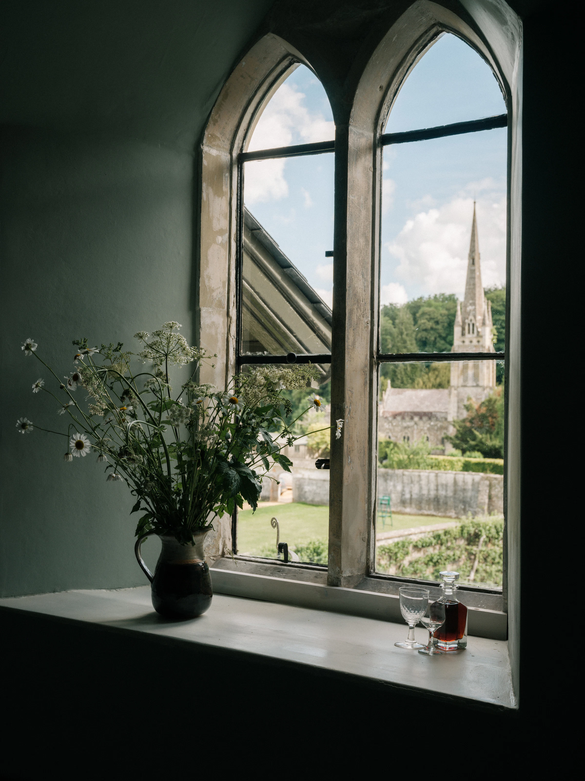 A window with a vase of wildflowers, two glasses, and a decanter, overlooking a church and garden outside.