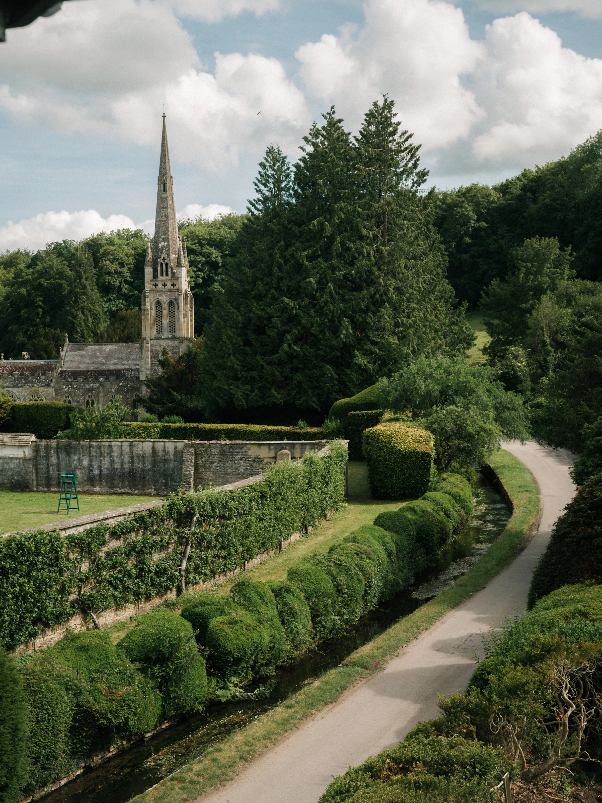 A winding country road passes by a church with a tall spire, surrounded by lush greenery and manicured hedges.
