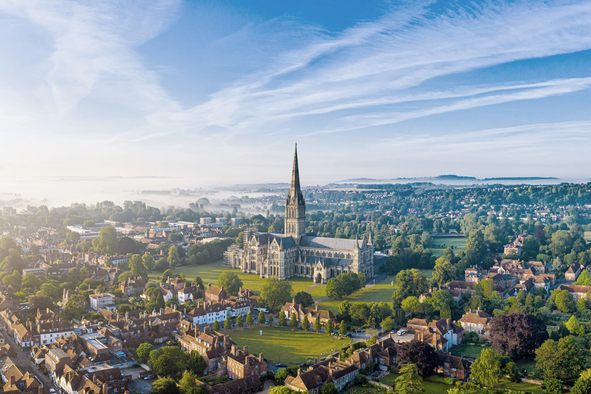 Aerial view of Salisbury Cathedral surrounded by trees, houses, and misty countryside under a blue sky with wispy clouds.