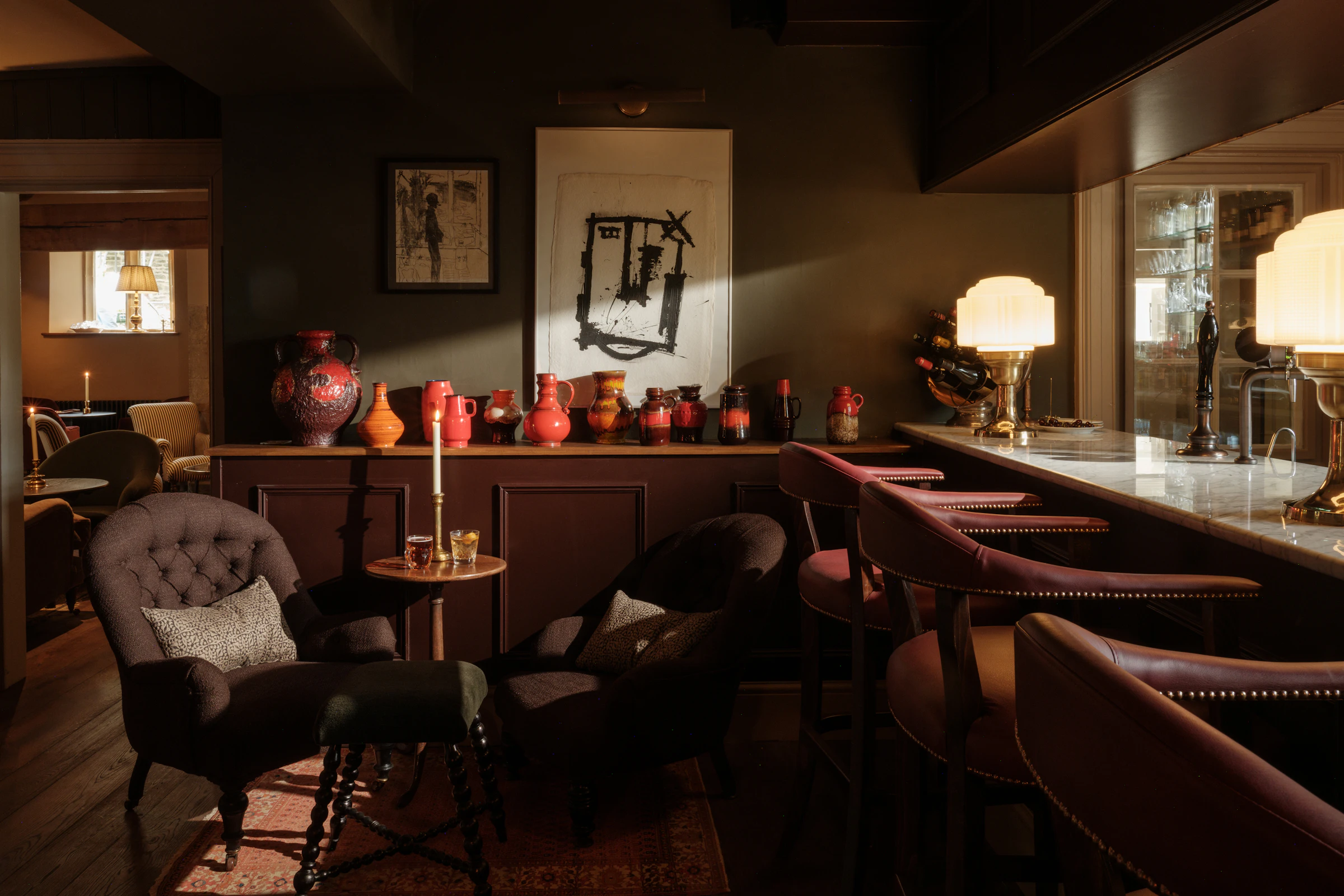 Dimly lit lounge with armchairs, bar stools, red vases, framed art, and warm table lamps on a marble counter.